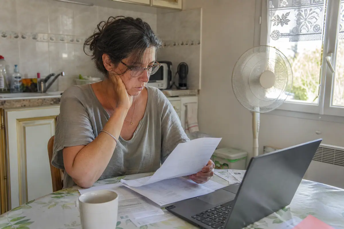 Une femme consulte sa facture d'électricité sur son ordinateur avec un ventilateur en marche à l'arrière plan