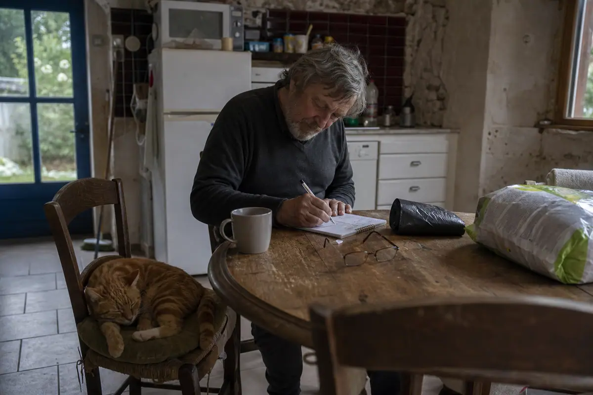 Un homme retraité calcule ses dépenses dans un carnet à côté d'un sac de litière et de son chat endormi