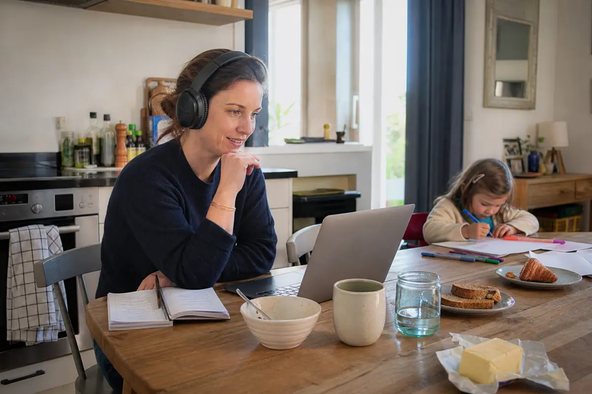 Une mère en télétravail participe à une visioconférence avec un casque pendant que sa fille dessine à côté