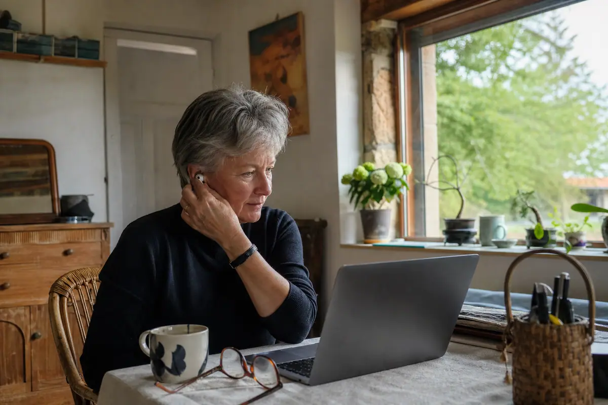 Une femme d'une cinquantaine d'années ajuste un écouteur sans fil pendant un appel vidéo depuis son bureau à la maison