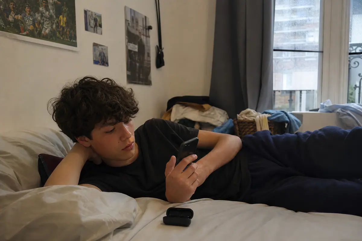 Un adolescent allongé sur son lit choisit de la musique sur son téléphone à côté d'un étui d'écouteurs sans fil
