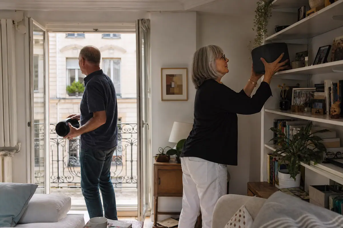 Un couple de retraités installe une enceinte sur une étagère pendant que l'homme emporte une enceinte portable vers le balcon