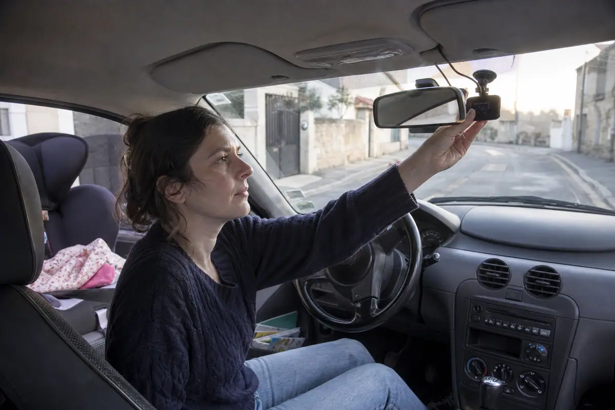 Une conductrice fixe une petite dashcam derrière le rétroviseur de sa voiture garée dans une rue résidentielle