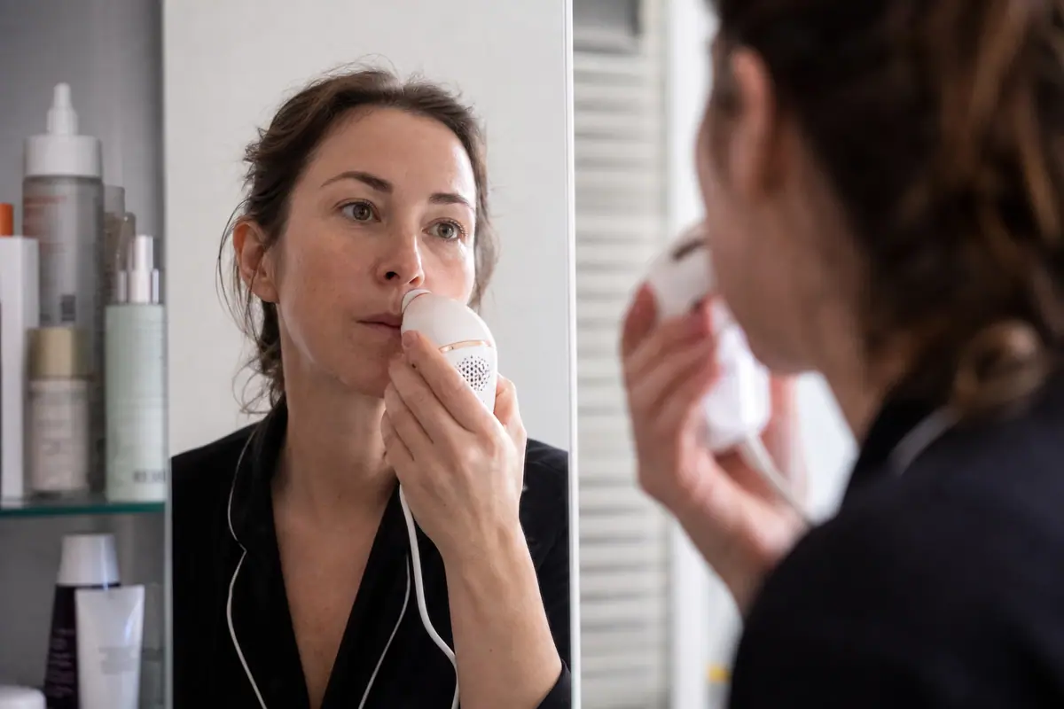 Une femme utilise un épilateur à lumière pulsée près de la lèvre supérieure devant un miroir de salle de bain