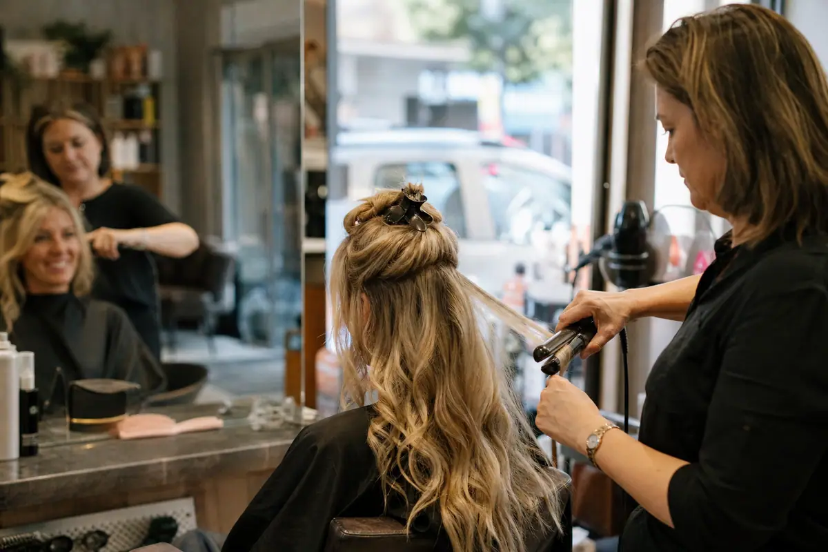 Une coiffeuse boucle les longs cheveux blonds d'une cliente avec un lisseur dans un salon de quartier