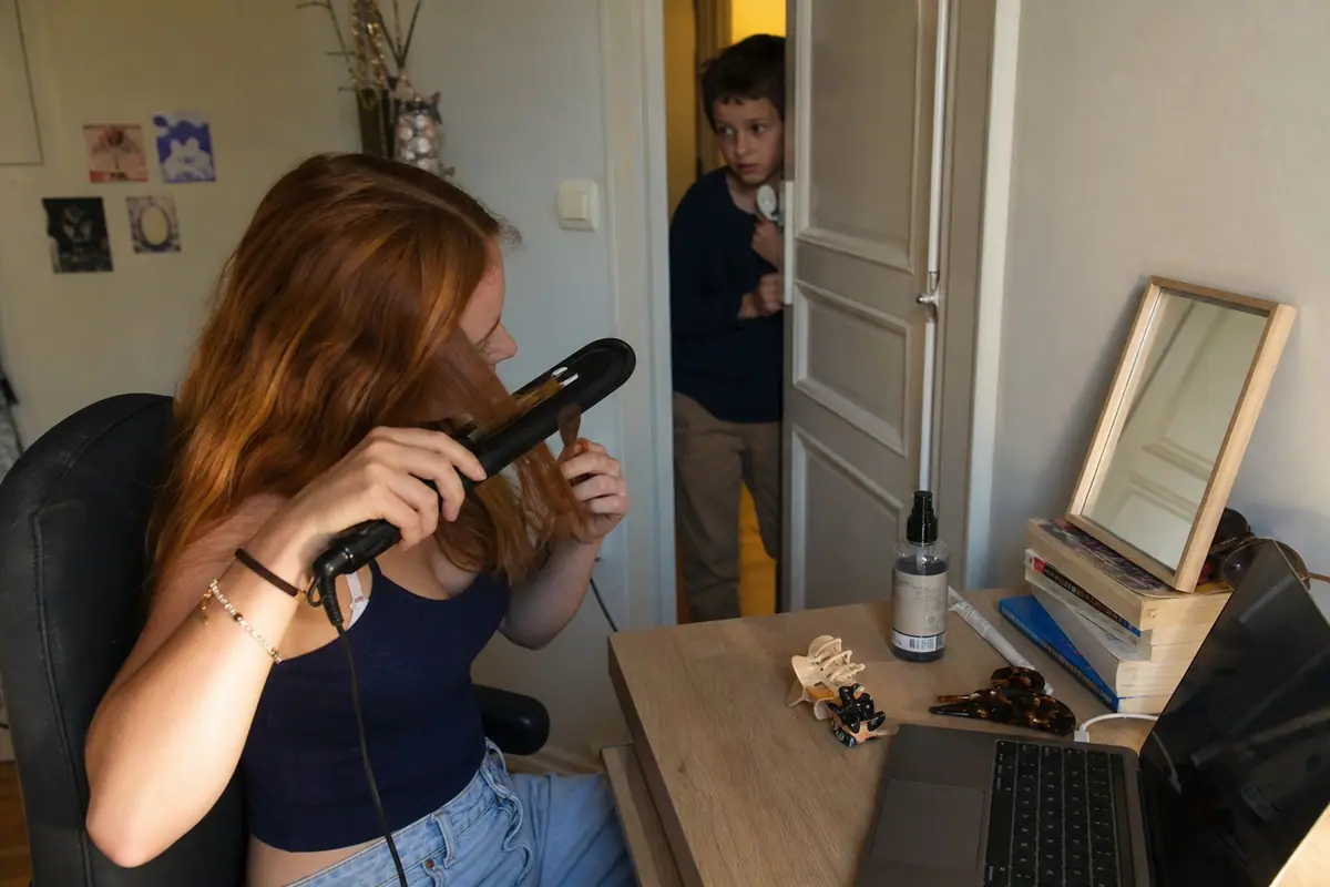 Une adolescente lisse une mèche de ses longs cheveux ondulés avec un lisseur devant un petit miroir