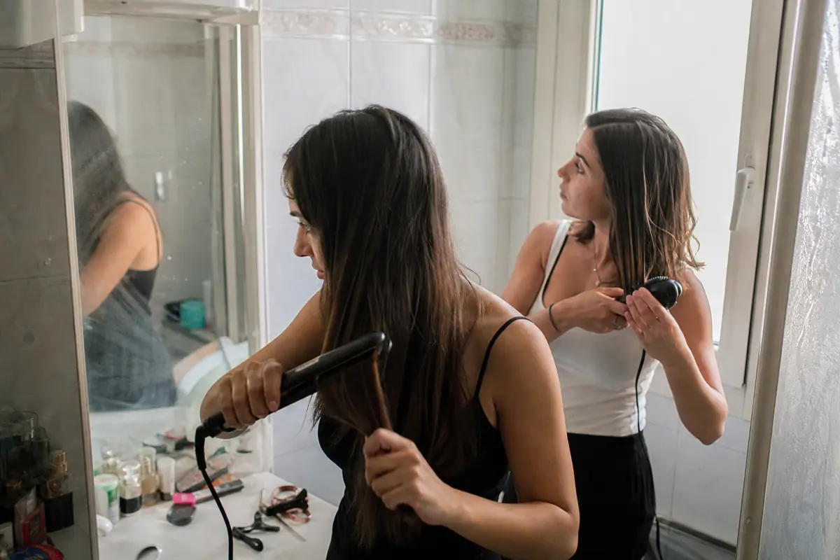 Deux sœurs se coiffent devant le miroir, l'une avec un lisseur, l'autre avec une brosse lissante chauffante
