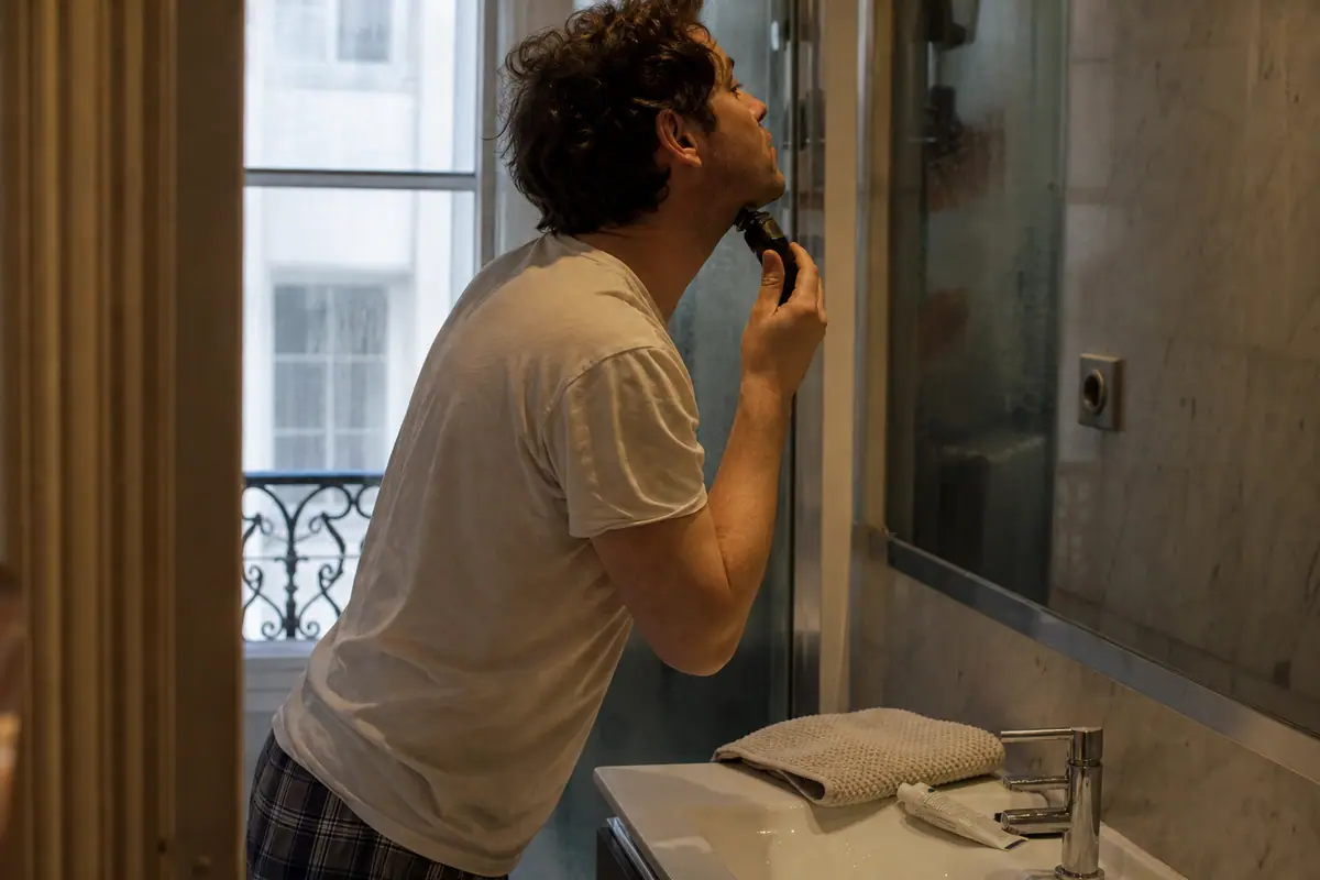 Un homme se rase le visage avec un rasoir électrique devant le miroir embué de sa salle de bain