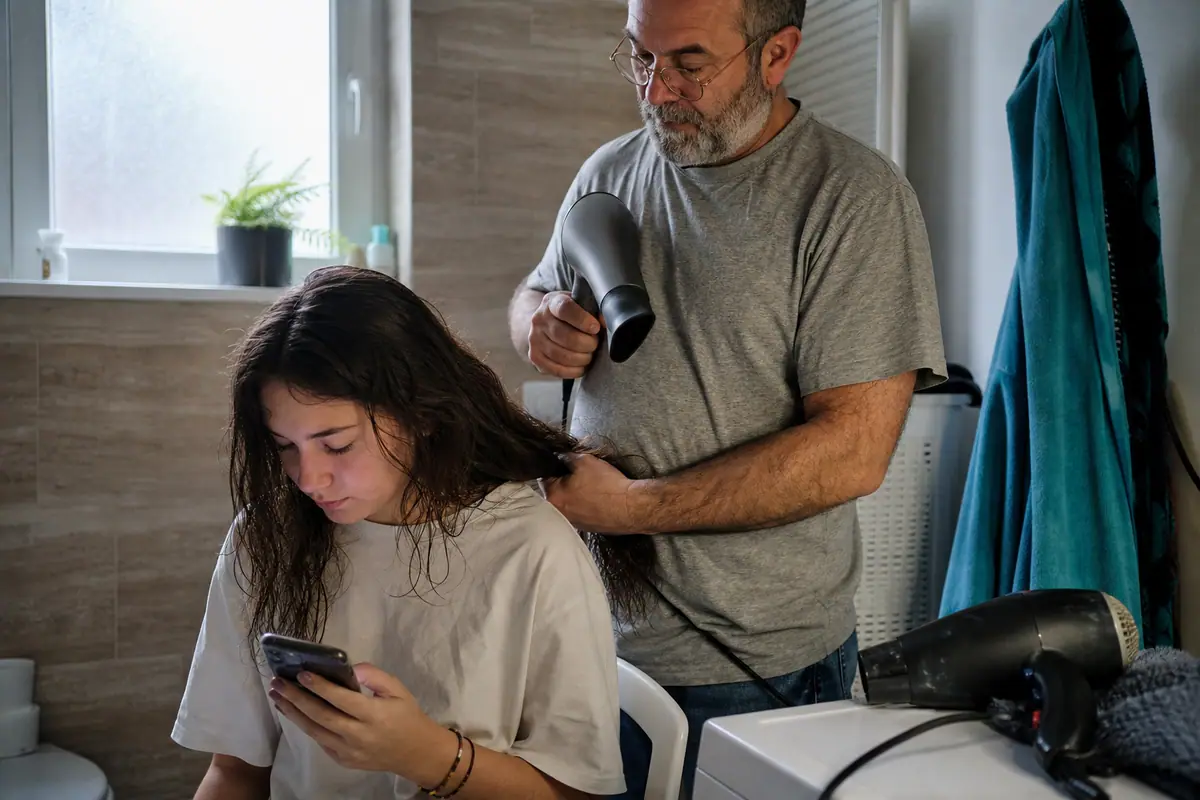 Un père sèche les cheveux de sa fille adolescente dans la salle de bains un matin d'école