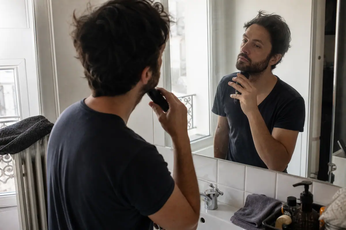 Un homme entretient sa barbe devant le miroir de sa salle de bain avec une tondeuse sans fil