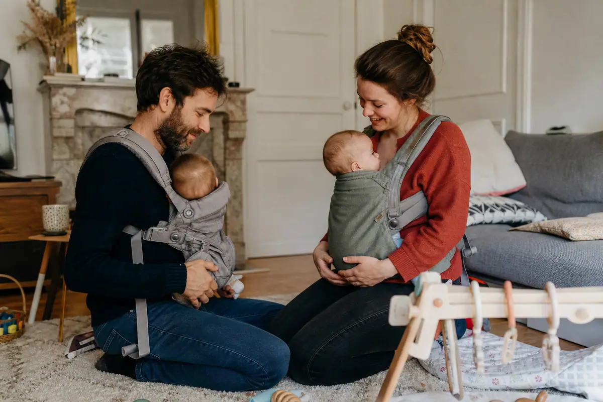 Deux parents comparent leurs porte-bébés physiologiques assis sur le tapis du salon avec leurs bébés
