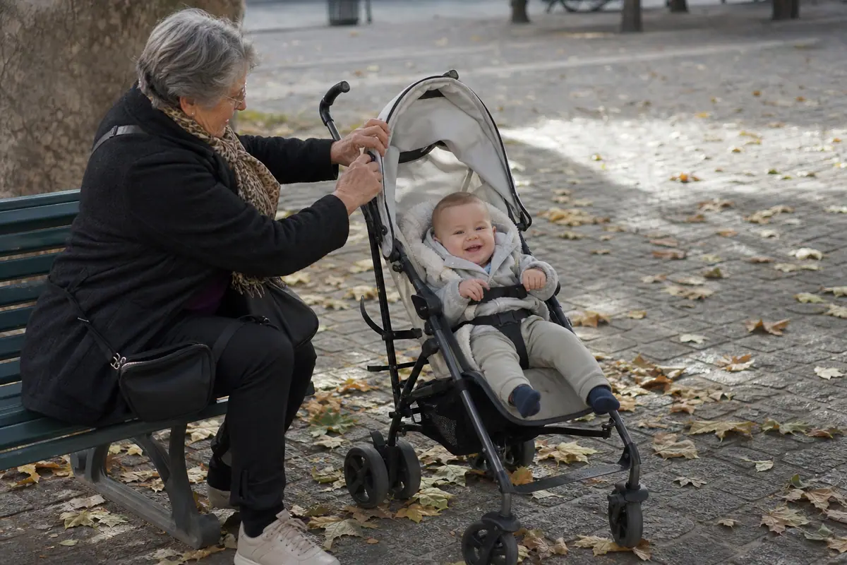 Une grand-mère règle le dossier d'une poussette canne où un bébé de sept mois est assis dans un square