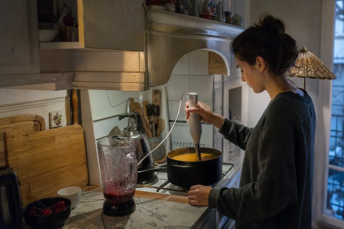 Une femme mixe une soupe directement dans la casserole avec un mixeur plongeant, un blender posé à côté sur le plan de travail