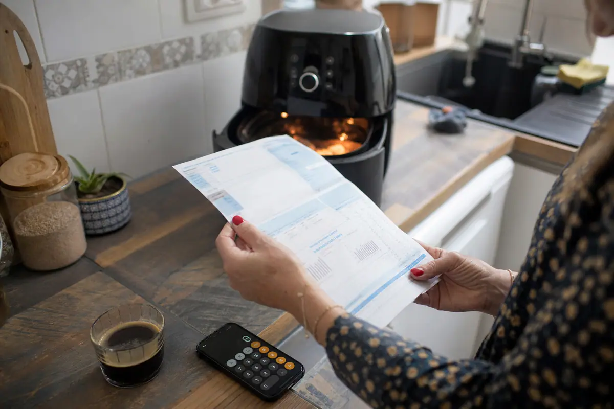 Une femme calcule sa consommation électrique avec sa facture à côté d'une friteuse sans huile en fonctionnement