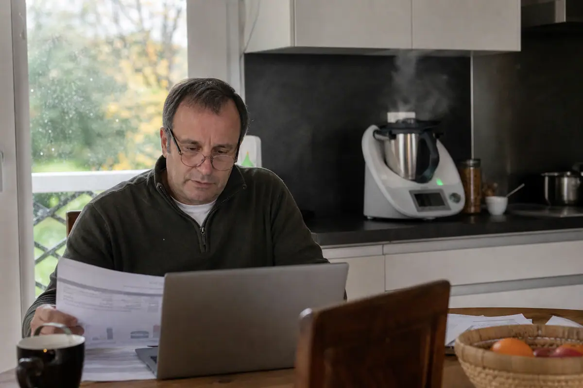 Un homme vérifie sa facture d'électricité sur son ordinateur pendant qu'un robot cuiseur mijote une soupe