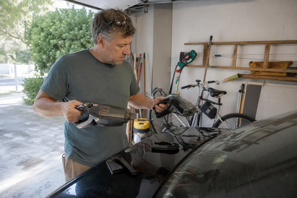 Un homme compare deux aspirateurs à main posés sur le coffre de sa voiture dans son garage