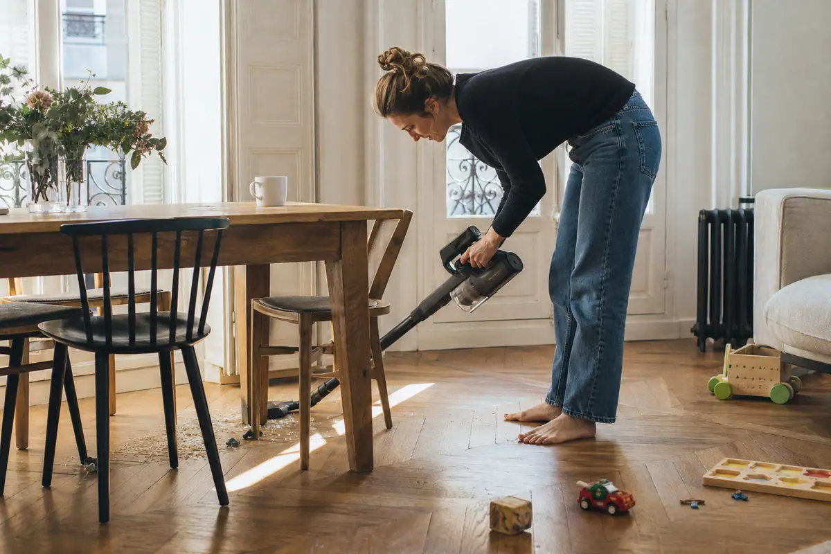 Une femme passe un aspirateur balai sans fil sous la table du salon, entre les jouets de son enfant