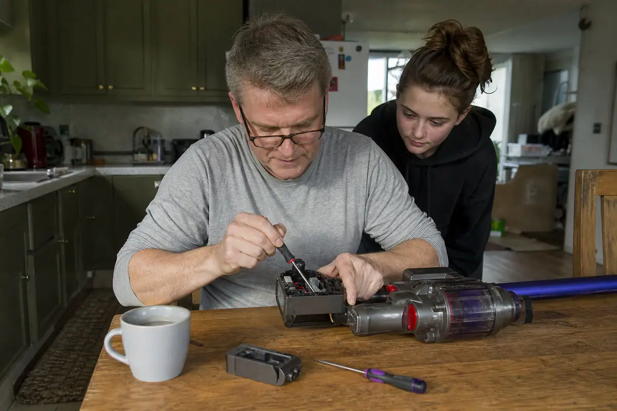 Un homme remplace la batterie de son aspirateur balai sur la table de la cuisine sous le regard de sa fille