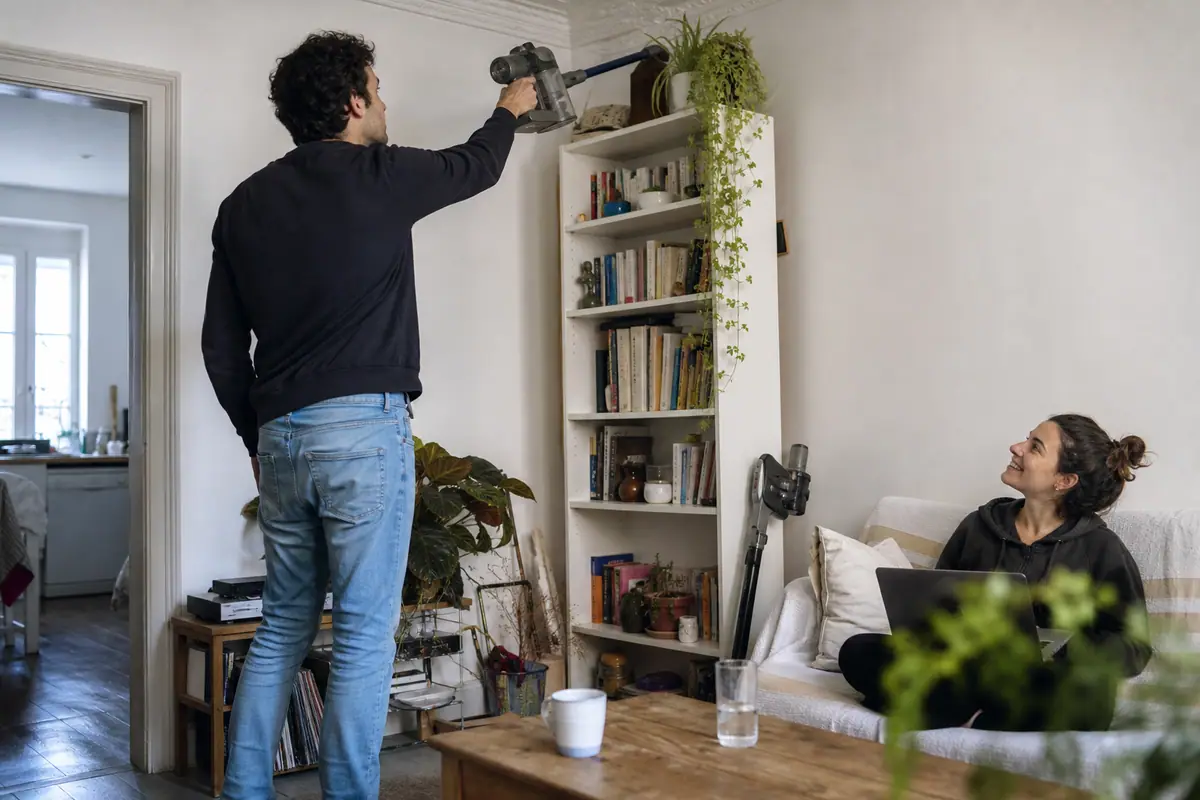 Un jeune homme aspire le dessus d'une bibliothèque avec un aspirateur balai léger dans un petit appartement parisien