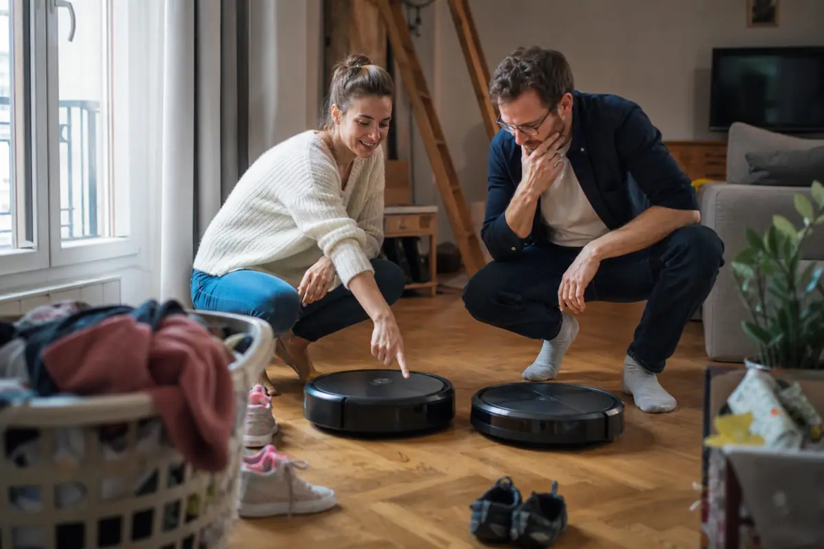 Un couple compare deux aspirateurs robots posés côte à côte sur le parquet du salon