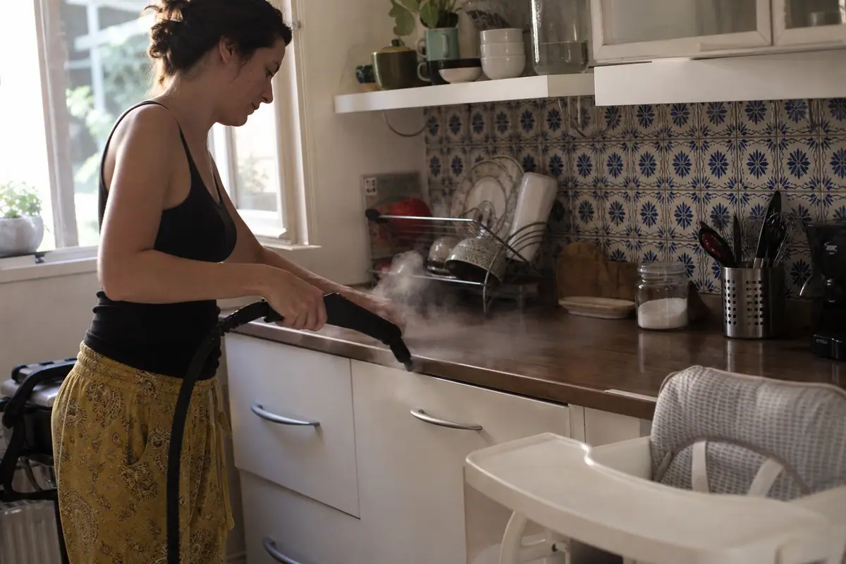 Une femme désinfecte son plan de travail de cuisine avec un nettoyeur vapeur près de la chaise haute de son bébé