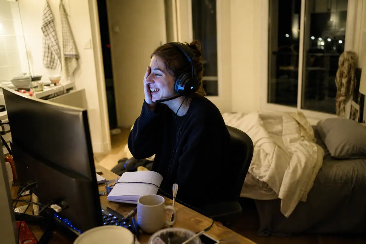 Une jeune femme coiffée d'un casque gaming rit en discutant avec ses amis devant son écran, le soir dans sa chambre