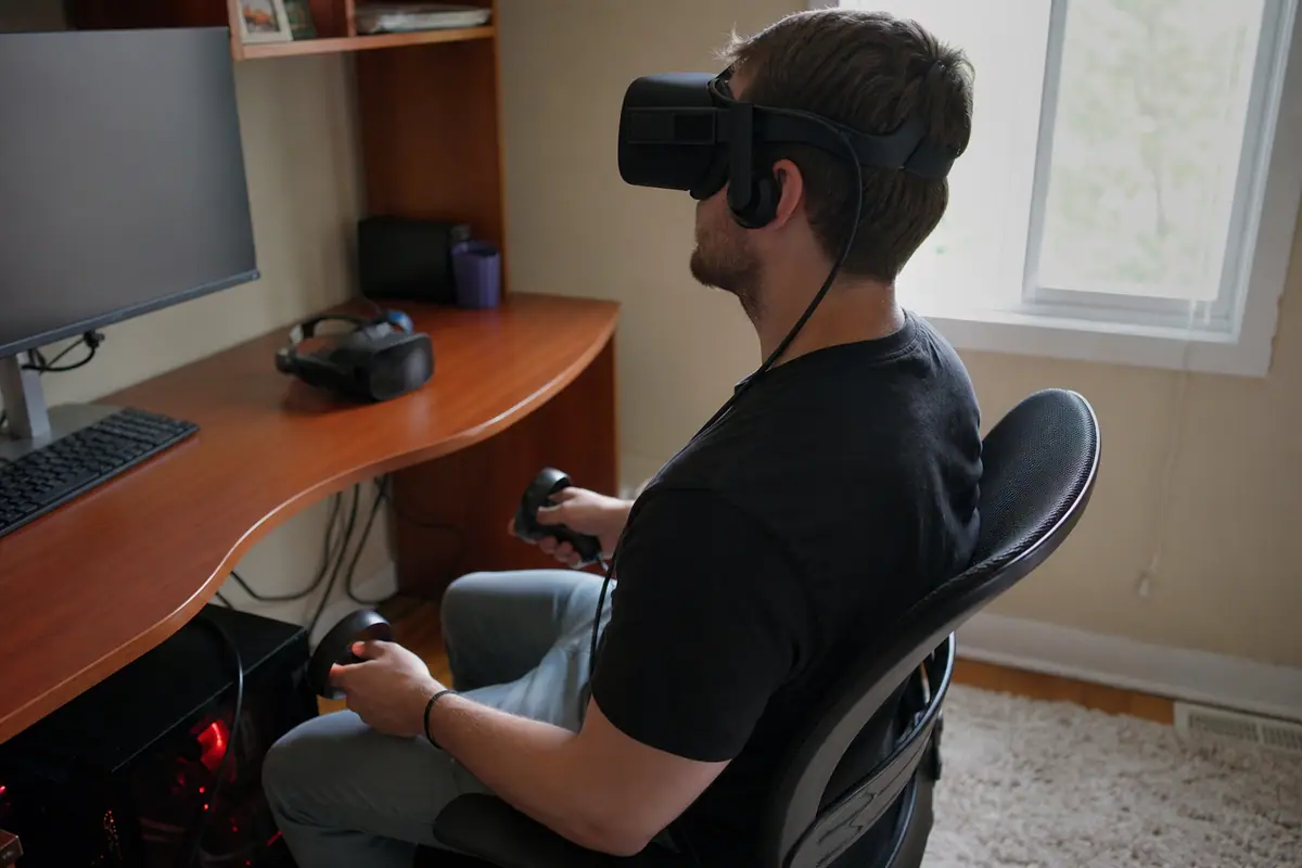 Un homme joue avec un casque de réalité virtuelle relié par câble à son ordinateur, un second casque autonome posé sur le bureau