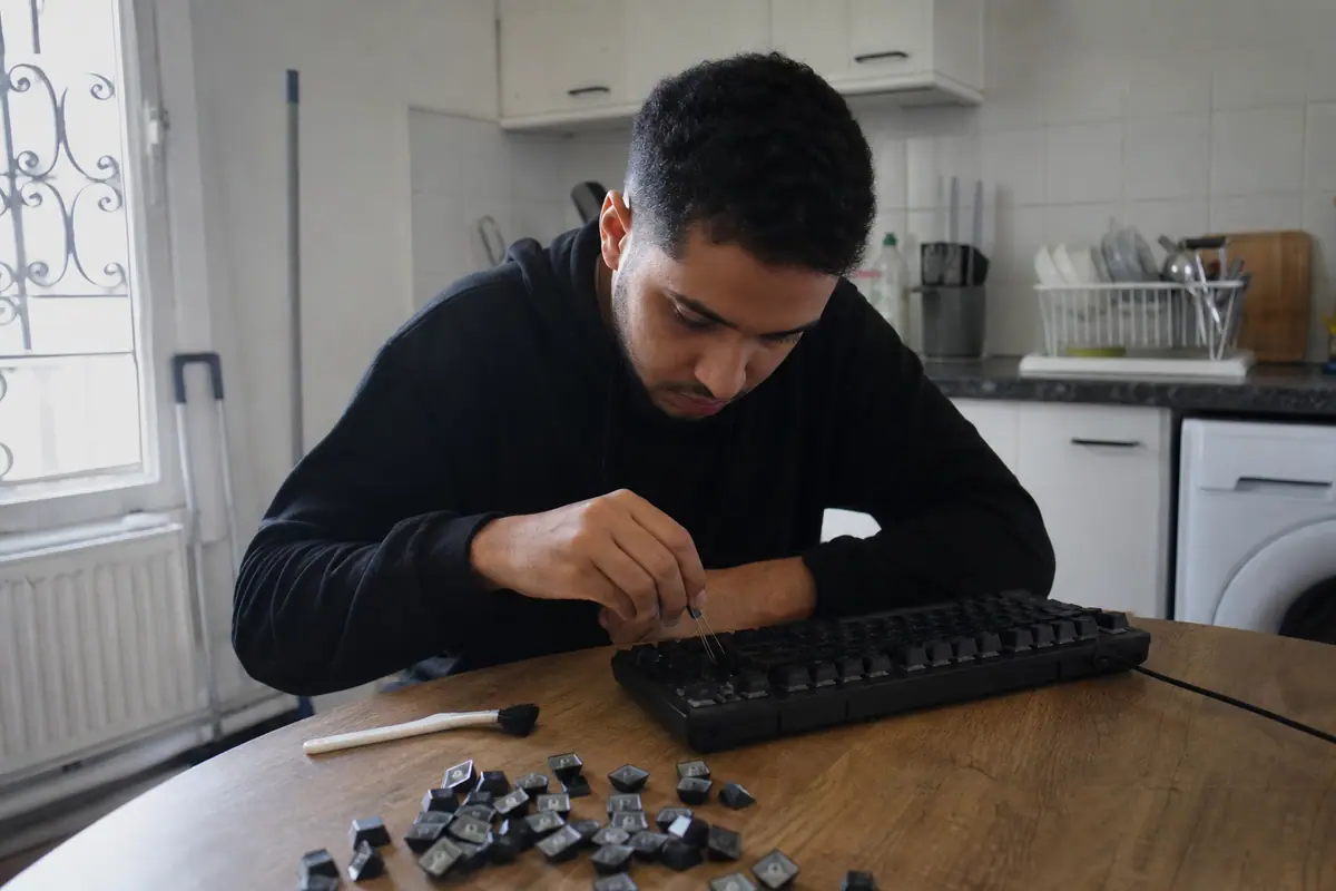 Un jeune homme retire un capuchon de touche de son clavier gaming mécanique avec un outil dédié, sur sa table de cuisine