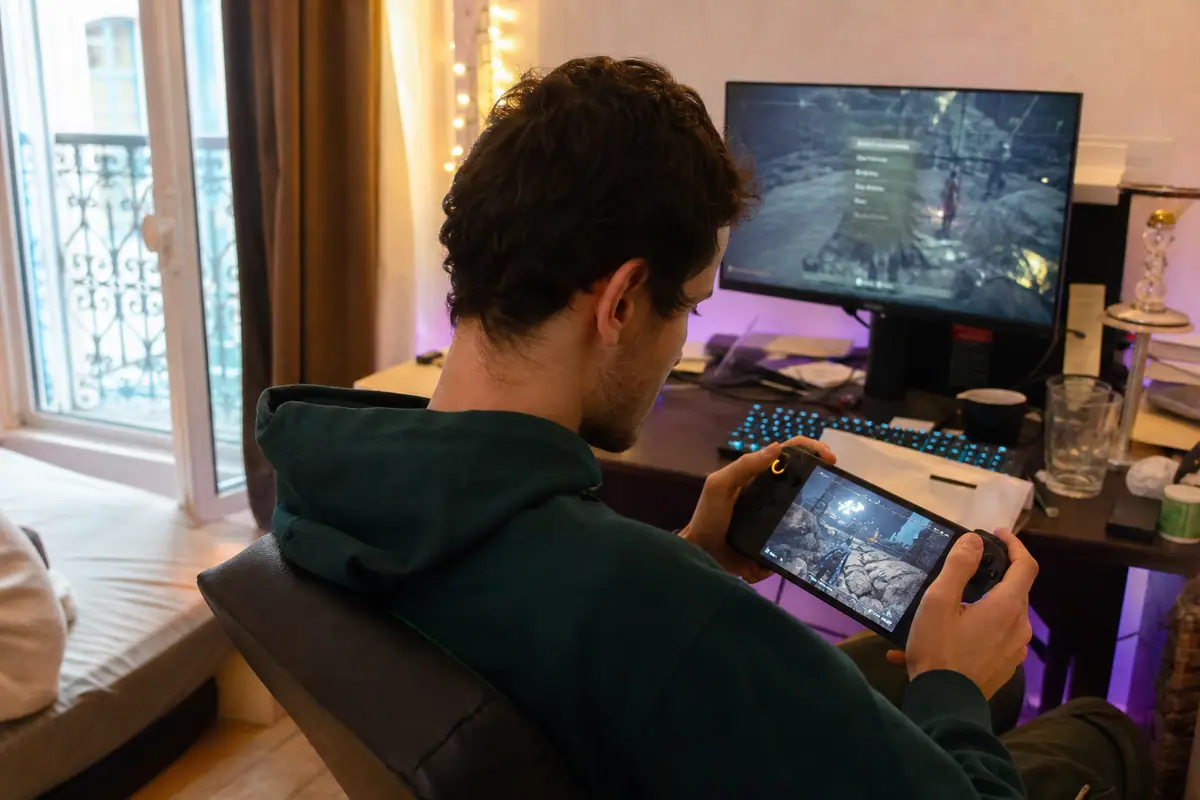 Un jeune homme joue sur une console portable PC à son bureau, un écran d'ordinateur allumé derrière lui