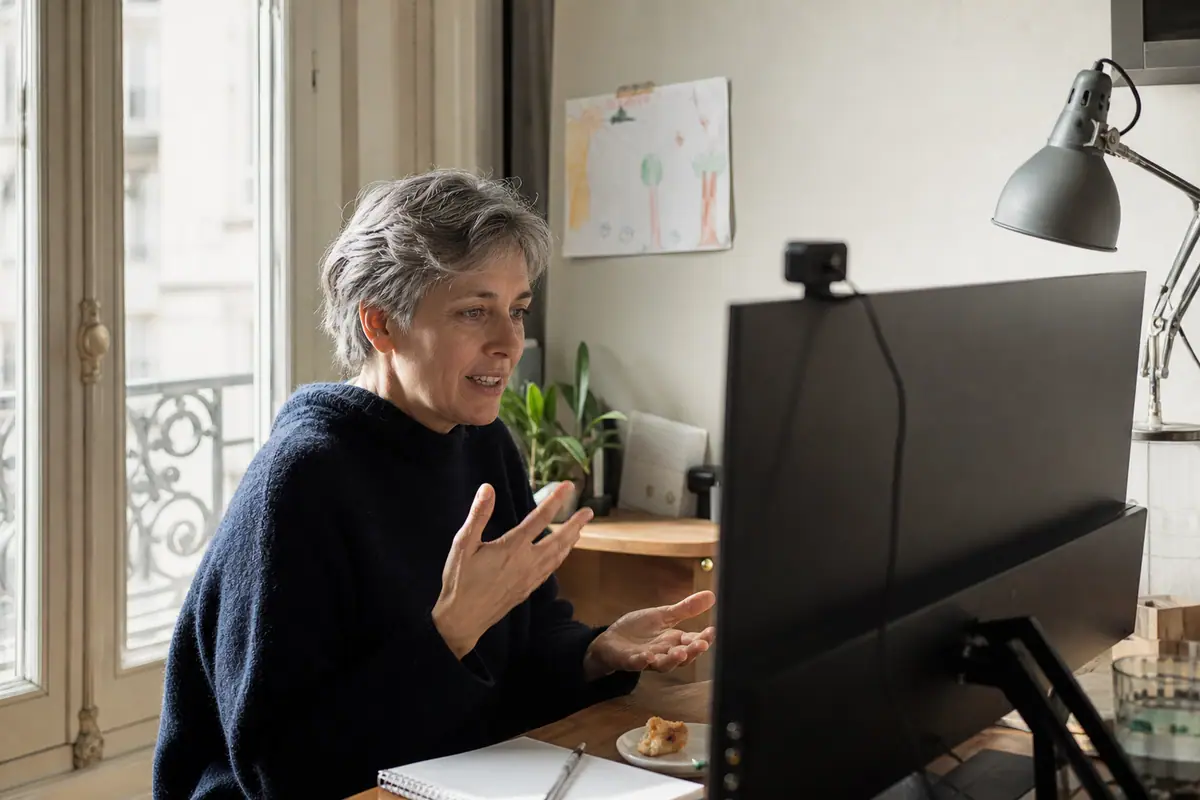Une femme participe à une visioconférence depuis son salon avec une webcam fixée sur son écran