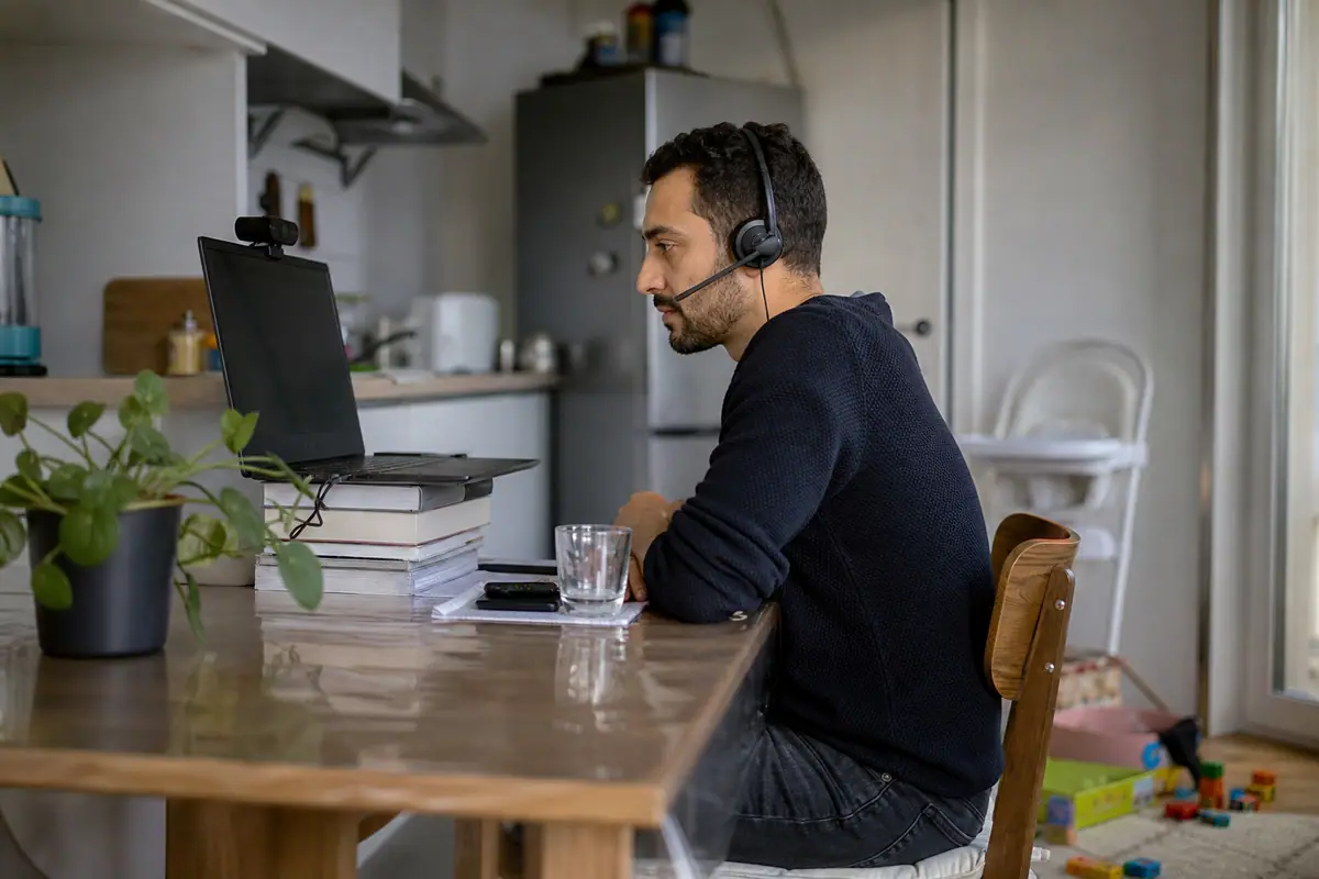 Un homme en télétravail participe à une réunion vidéo depuis sa cuisine, son ordinateur surélevé sur des livres avec une webcam externe