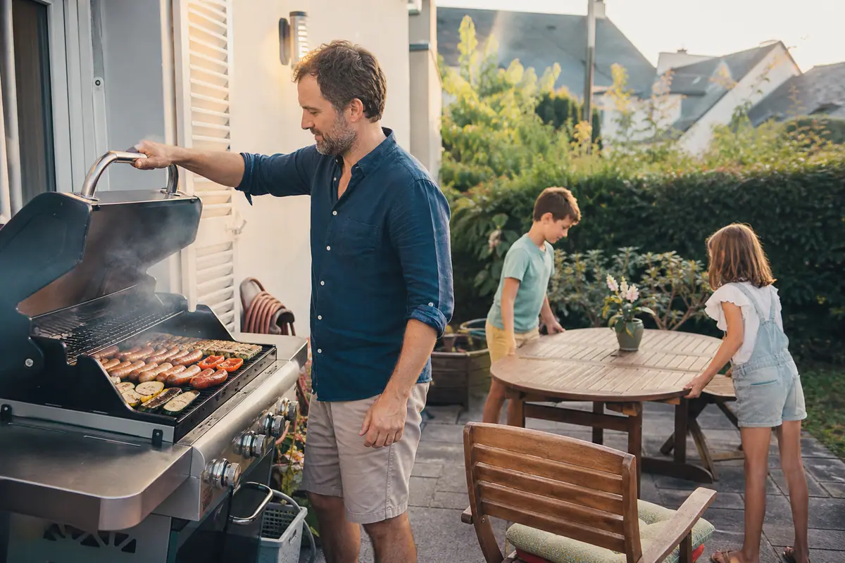 Un père soulève le couvercle d'un barbecue à gaz sur sa terrasse pendant que ses enfants mettent la table à proximité
