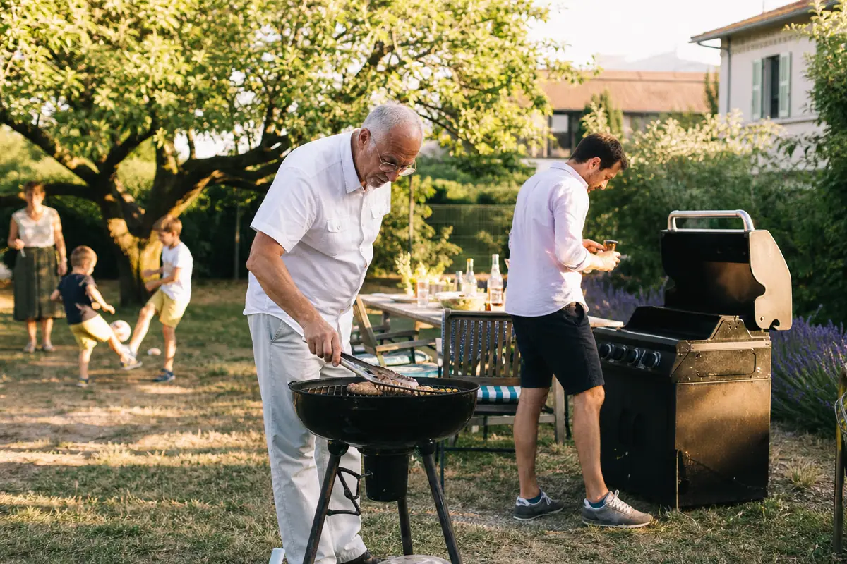 Un grand père surveille un barbecue au charbon pendant que son fils allume un barbecue à gaz dans le même jardin