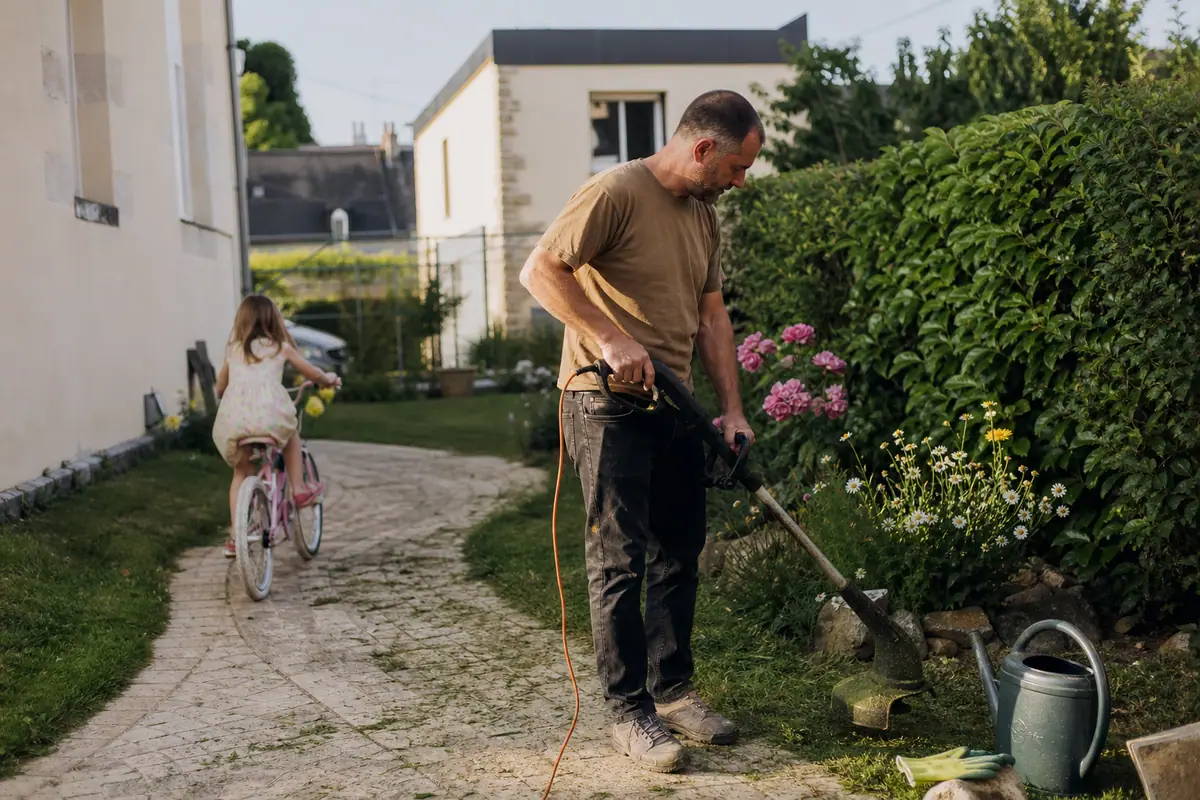 Un homme finit les bordures de sa pelouse au coupe-bordure pendant que sa fille joue à vélo dans le jardin