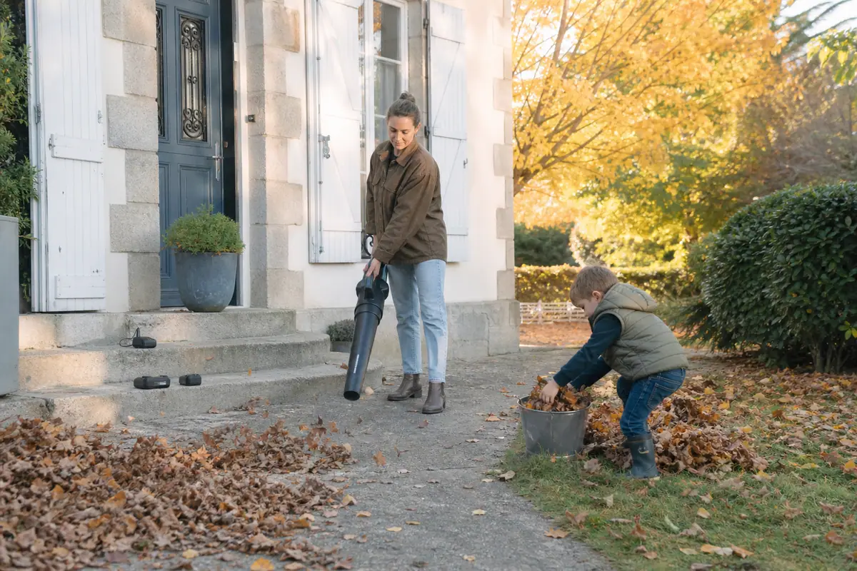 Une femme utilise un souffleur de feuilles sans fil dans son jardin pendant que son fils ramasse les feuilles dans un seau