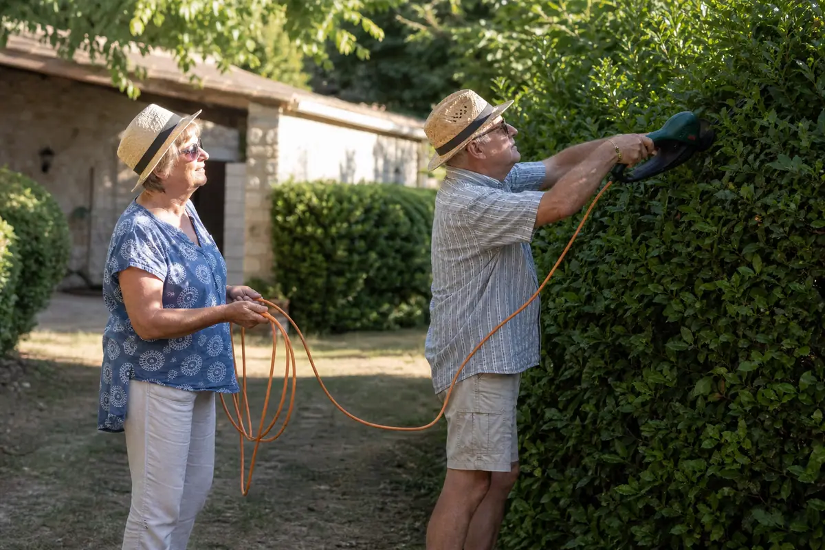 Un homme taille une haie avec un taille-haie filaire pendant que sa femme tient la rallonge à distance de la lame