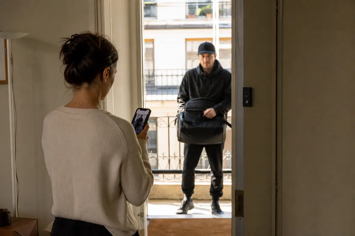 Une femme consulte la notification de sa sonnette connectée sur son smartphone pendant qu'un livreur attend devant la porte