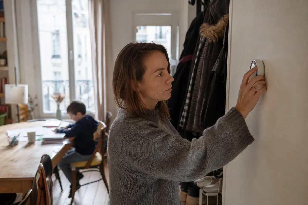 Une femme règle un thermostat connecté mural dans le couloir de son appartement en hiver