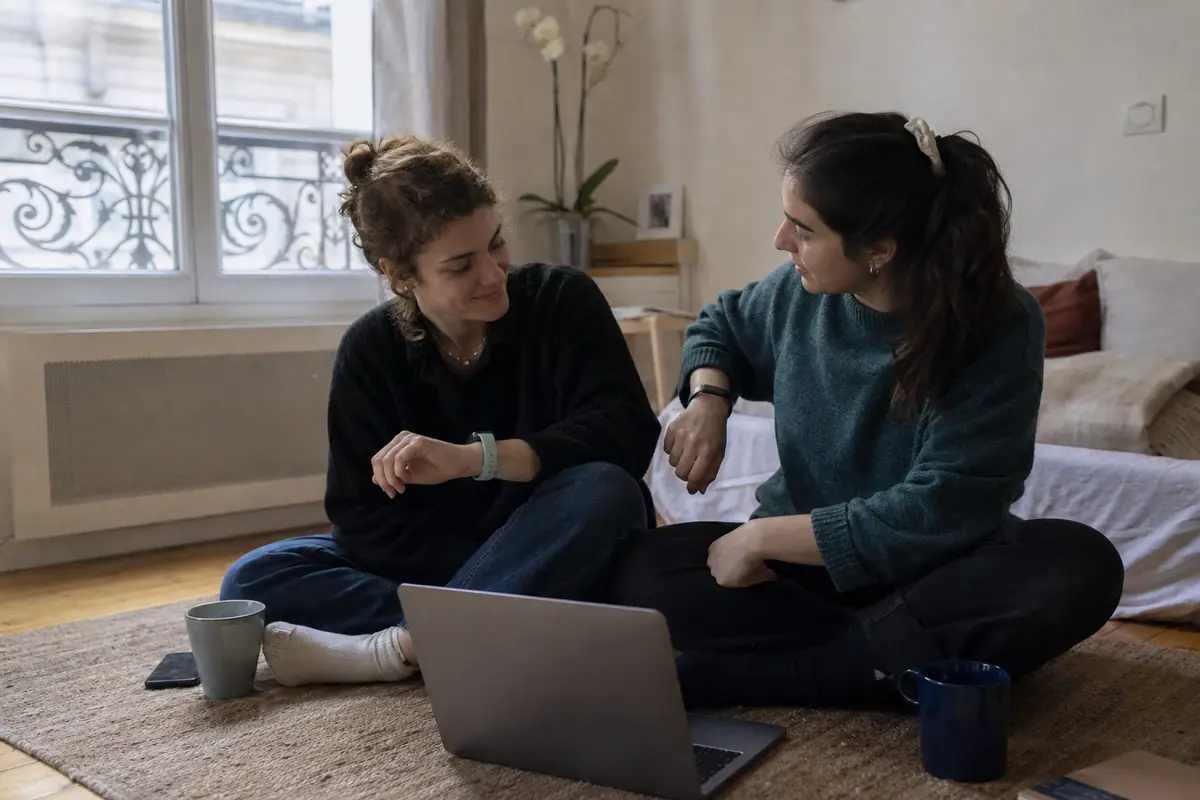 Deux colocataires comparent leurs bracelets connectés assises sur le tapis de leur appartement