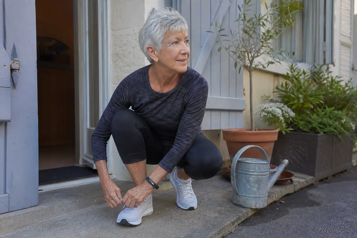 Une femme d'une soixantaine d'années lace ses chaussures de course sur le pas de sa porte, une montre connectée fine au poignet