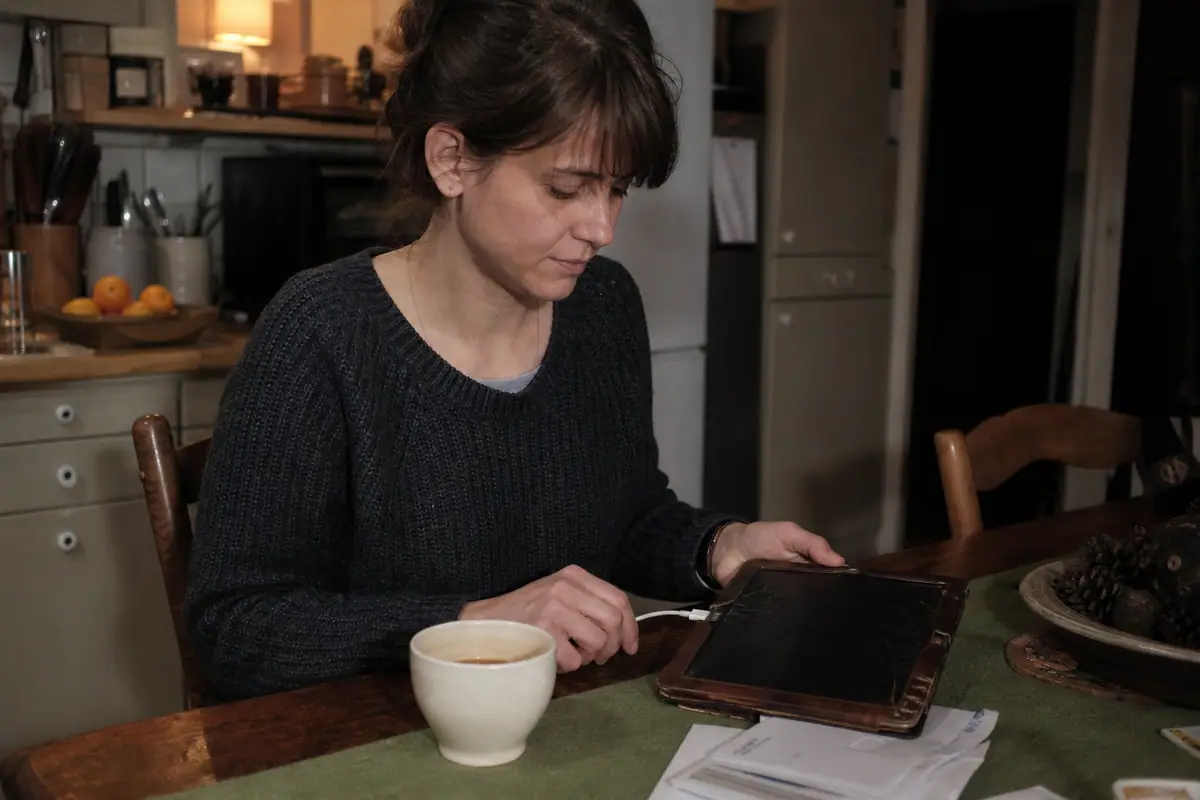 Une femme branche une vieille tablette à la coque usée sur son chargeur dans sa cuisine le soir