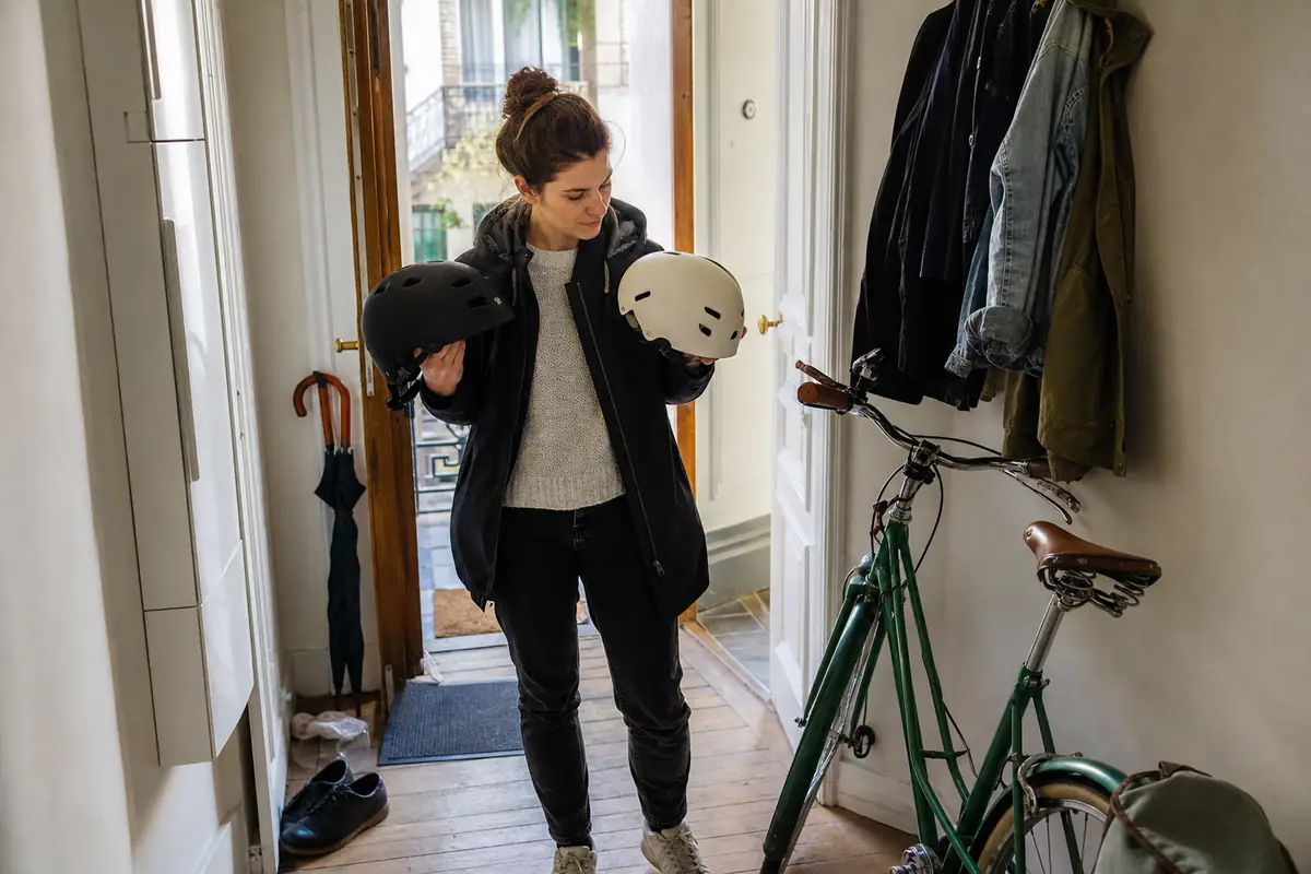 Une femme compare deux casques de vélo dans l'entrée de son appartement avant de partir travailler
