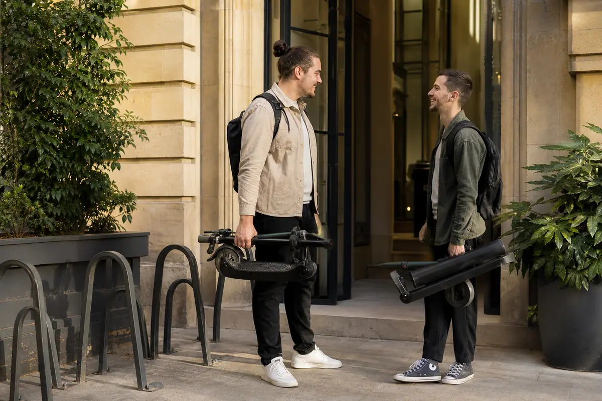 Deux collègues comparent leurs trottinettes électriques pliées devant l'entrée de leur bureau
