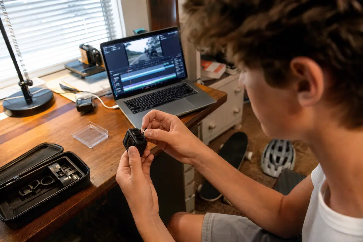 Un adolescent insère une carte mémoire dans sa caméra sport à son bureau, un casque de vélo et un skateboard à ses pieds
