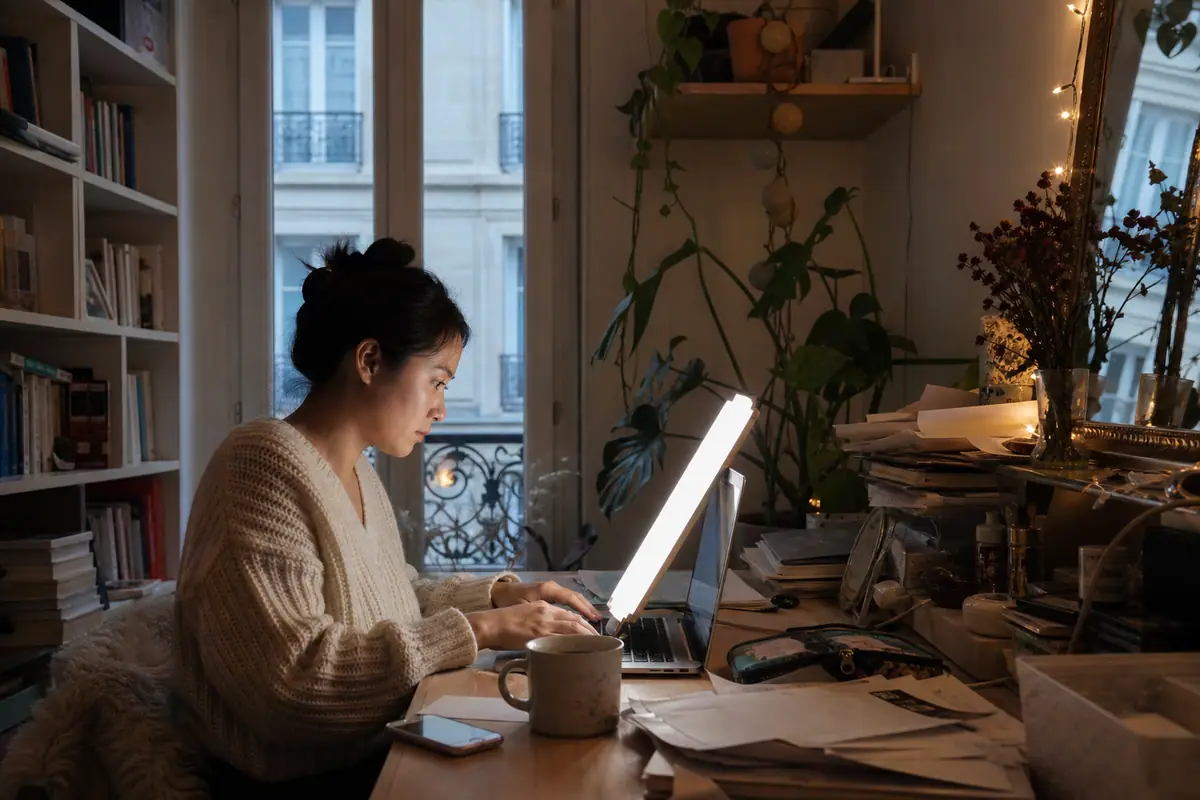 Une jeune femme travaille à son bureau à domicile avec une lampe de luminothérapie fine appuyée contre son écran