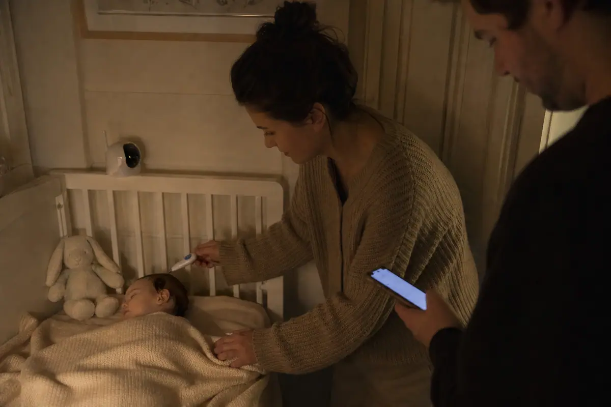 Une mère mesure la température de son bébé endormi dans son berceau pendant que son partenaire note le résultat sur son téléphone