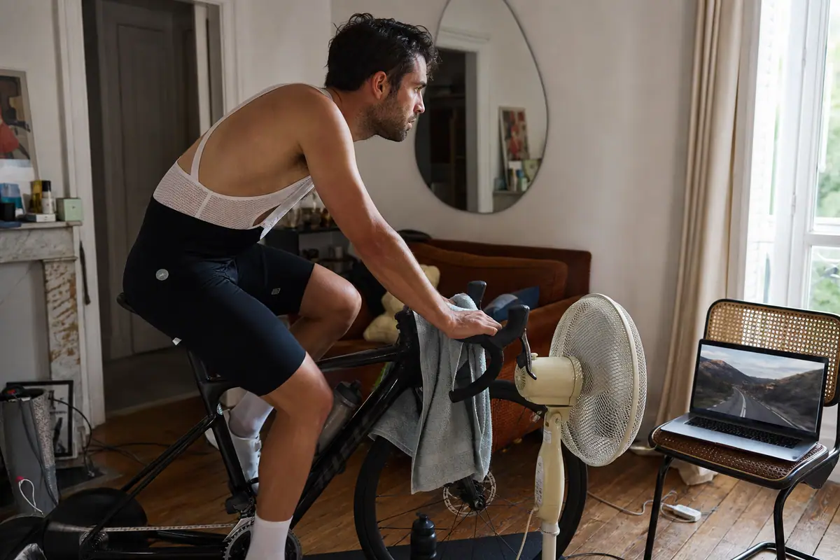 Un homme pédale sur son vélo monté sur un home trainer dans le coin de son salon, un ventilateur à ses côtés