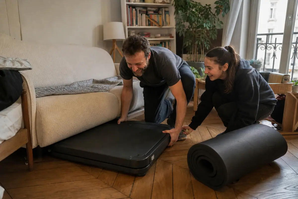 Un père et sa fille glissent un tapis de course replié à plat sous le canapé de leur salon