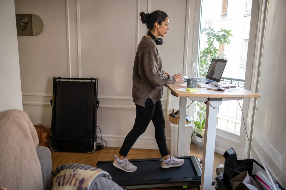 Une jeune femme marche sur un tapis de marche pliable sous son bureau debout pendant une visioconférence