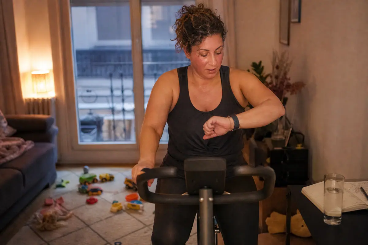 Une femme d'une quarantaine d'années pédale sur un vélo d'appartement en regardant sa montre de sport
